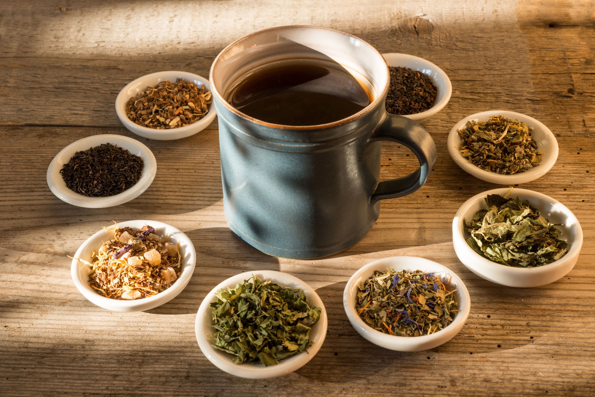 Different kinds of tea or herbs in bowls on an old table with cup of tea in sunset